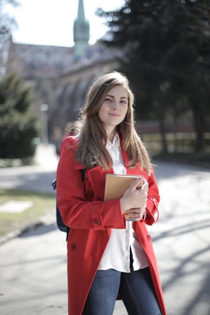 pexels-photo-3793238-3793238 Young woman in a red coat smiling while holding a book outside on a sunny day.